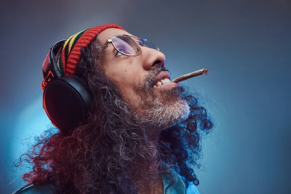 Studioporträt des afrikanischen rastafarischen Männchens genießt Musik im Kopfhörer und raucht Unkraut. — Stockfoto