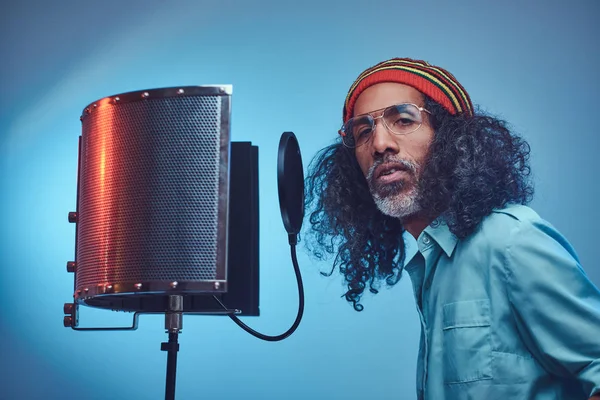 Αφρικανός Rastafarian τραγουδιστής άντρας φορώντας ένα μπλε πουκάμισο και beanie τραγούδι συναισθηματικής γραφής στο στούντιο ηχογράφησης. — Φωτογραφία Αρχείου