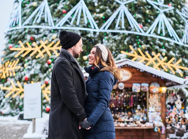 Casal jovem vestindo roupas quentes de mãos dadas e olhar uns para os outros, de pé perto de uma árvore de Natal da cidade, gostando de passar o tempo juntos . — Fotografia de Stock