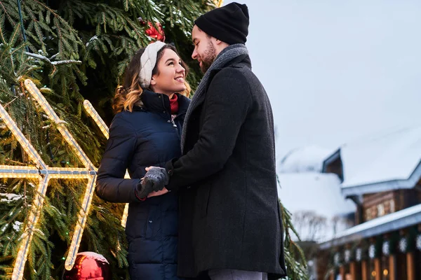 Pareja joven con ropa de abrigo cogida de la mano y mirándose, de pie cerca de un árbol de Navidad de la ciudad, disfrutando pasar tiempo juntos . — Foto de Stock