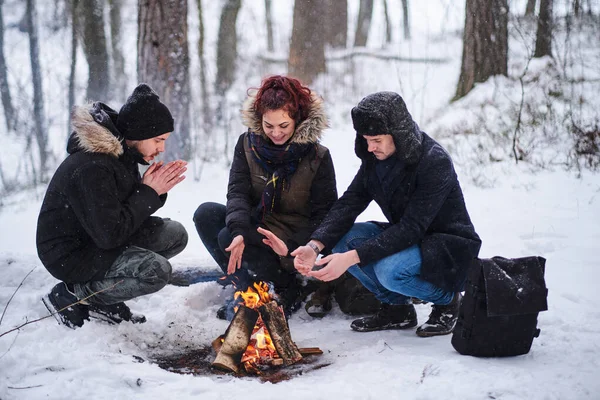 Amigos de viajantes se aquecem em um incêndio na floresta nevada — Fotografia de Stock