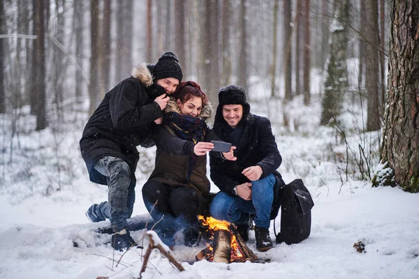 Turisták ülnek tábortűz mellett, hogy szelfi fotó okostelefon a hideg havas erdőben — Stock Fotó