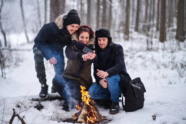Tourists sit next to bonfire taking selfie photo smartphone in the cold snowy forest