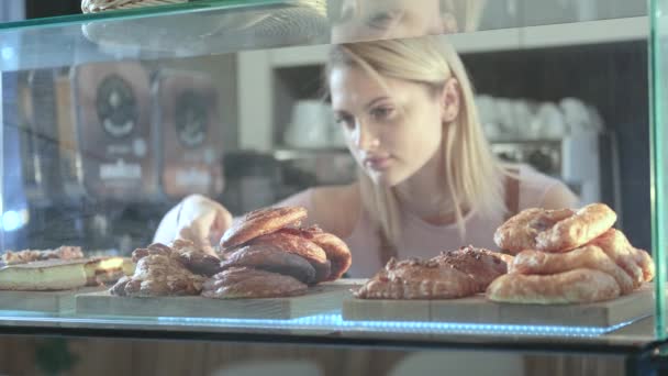 Gyönyörű fiatal női eladó, aki a cukrászdában dolgozik. Friss péksütemények közelsége vitrinen — Stock videók