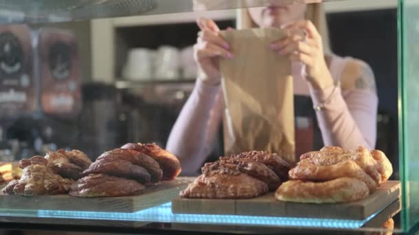 Bella giovane venditrice che lavora nel negozio di dolciumi. Primo piano di pasticcini freschi. Venditore confeziona panino fresco in un sacchetto di carta — Video Stock