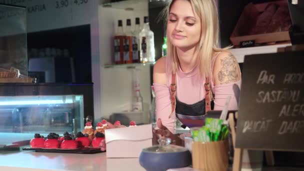 Beautiful young girl seller shows mouthwatering cakes standing behind the counter — ストック動画