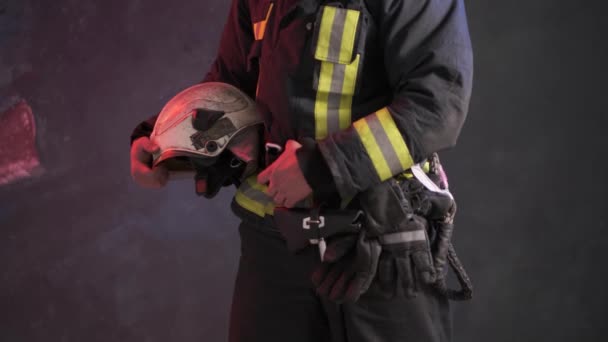 Vista da vicino di un coraggioso vigile del fuoco in uniforme speciale tiene casco protettivo in piedi contro una parete grigia — Video Stock