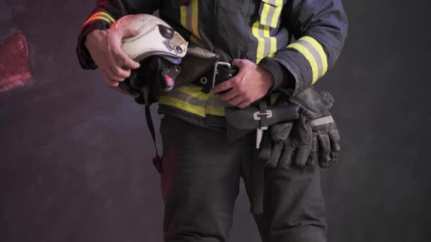 Vista de cerca de un valiente bombero con uniforme especial sostiene el casco protector de pie contra una pared gris — Vídeos de Stock