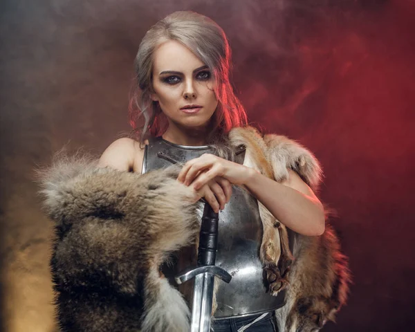 Portrait of a beautiful warrior woman holding a sword wearing steel cuirass and fur. Fantasy fashion. Studio photography on a dark background with red light. Cosplayer as Ciri from The Witcher. — Stock Photo, Image