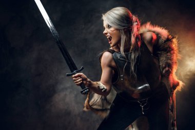 Kürklü ve kürklü fantezi kadın şövalye, bir kılıç kınını savaşa hazır tutuyor. Fantezi modası. Ciri rolünde Cosplayer. Stüdyo fotoğrafçılık