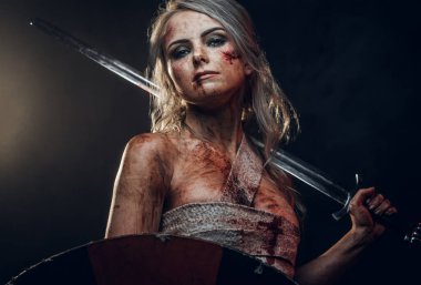 Kan ve çamurla lekelenmiş paçavra giysili kadın savaşçı. Kılıç ve kalkan taşıyor. Karanlık bir arka planda stüdyo fotoğrafı. Ciri rolünde Cosplayer Cadıdan