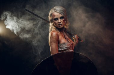 Fantezi kadın savaşçı, kana bulanmış bez giyip kılıçla ve kalkanla poz veriyor. Karanlık bir arka planda dumanlı stüdyo fotoğrafı. Ciri rolünde Cosplayer Cadıdan