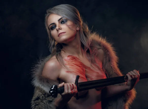 Portrét krásné nahé ženy s krví potřísněnou kůží, držící dlouhý meč a pózující před kamerou, oblečené pouze v kožichu. Fantazie. Cosplayer jako Ciri z The Witcher — Stock fotografie