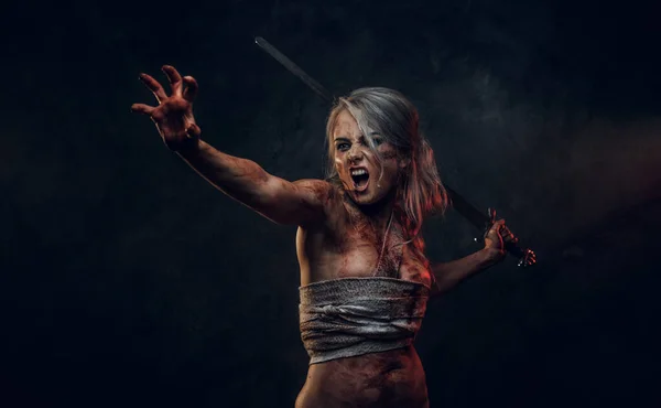 Žena, bojovnice v hadrech potřísněných krví a blátem, drží meč a se zuřivým výkřikem se řítí do boje. Studio fotografie na tmavém pozadí s kouřem. Cosplayer jako Ciri z The Witcher — Stock fotografie