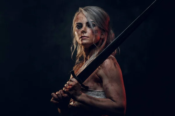 Meztelen női harcos rongyot visel, vérrel és sárral festve, karddal pózolva. Stúdió fotó sötét háttérrel. Cosplayer mint Ciri a Boszorkányból — Stock Fotó