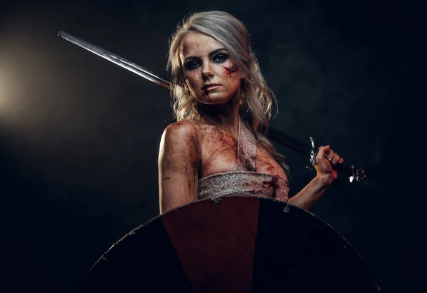 Woman warrior wearing rag cloth stained with blood and mud, holding sword and shield. Studio photo on a dark background. Cosplayer as Ciri from The Witcher — Stock Photo, Image