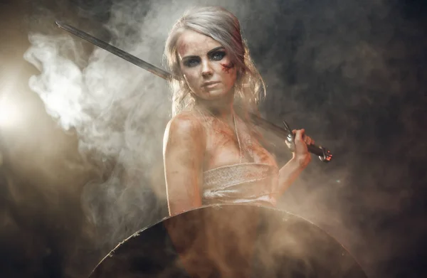 Fantasy woman warrior wearing rag cloth stained with blood and mud posing with a sword and shield. Studio photo on a dark background with smoke. Cosplayer as Ciri from The Witcher — 스톡 사진