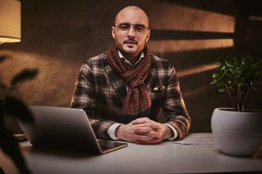 Bald european well-dressed serious businessman sitting in the office at a table with notebook, wearing stylish jacket and a scarf