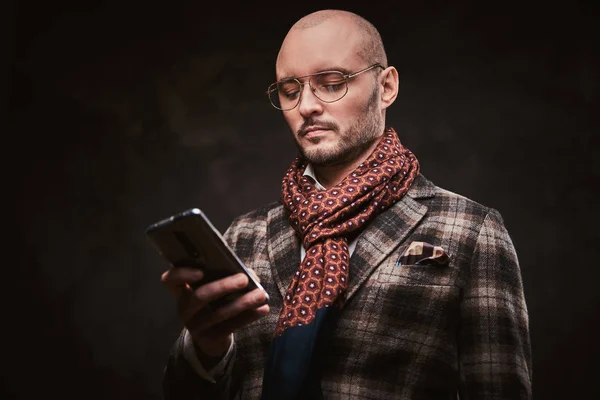 Úspěšný stylový podnikatel pózující ve studiu v kostkované bundě, brýlích a šále při držení smartphonu — Stock fotografie