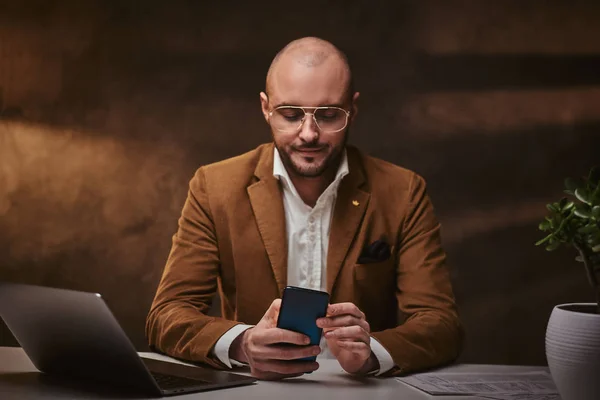 Calvo europeo bien vestido serio hombre de negocios sentado en la oficina en una mesa con portátil y teléfono inteligente, con elegante chaqueta de terciopelo y gafas —  Fotos de Stock