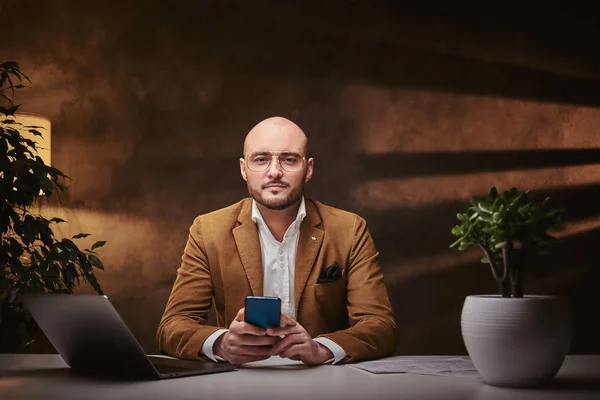 Καραφλός Ευρωπαίος καλοντυμένος σοβαρός επιχειρηματίας κάθεται στο γραφείο σε ένα τραπέζι με notebook και smartphone, φορώντας κομψό βελούδινο σακάκι και γυαλιά — Φωτογραφία Αρχείου