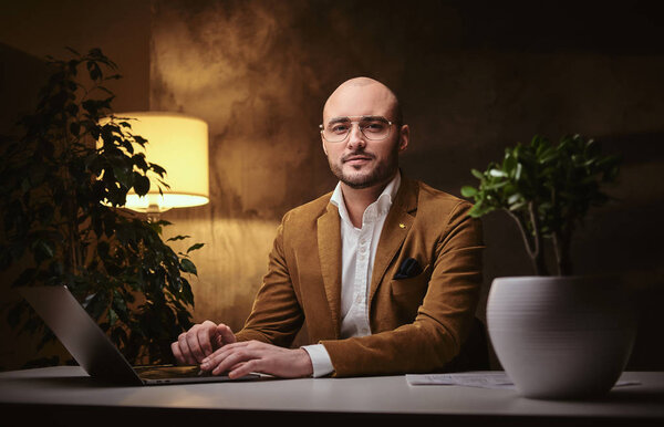 Лысый европейский хорошо одетый серьезный бизнесмен сидит в офисе за столом с ноутбуком, одетый в стильную бархатную куртку и очки
