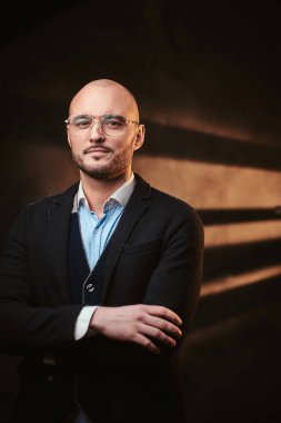 Siyah ceketli, mavi gömlekli ve gözlüklü, karanlık bir stüdyoda poz veren yakışıklı kel iş adamı..