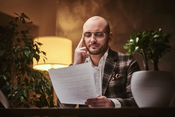 Glatzköpfiger, gut gekleideter Geschäftsmann sitzt im Büro an einem Tisch mit Notizbuch und Papieren und trägt eine stylische Jacke — Stockfoto