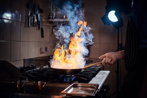 Cooker wearing gloves and apron frying flambe on a pan in a dark restaurant kitchen — Stock Photo, Image