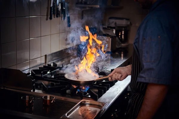 Chef wearing gloves and apron frying flambe on a pan in a dark restaurant kitchen — Stock Photo, Image