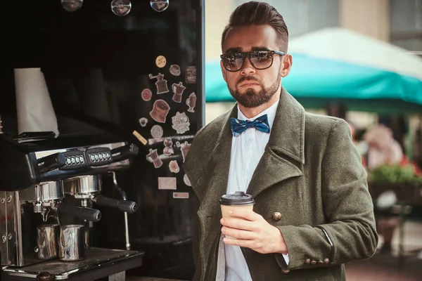 Elegante joven cliente masculino parado al aire libre en una ciudad, sosteniendo una taza de papel de café por la mañana — Foto de Stock