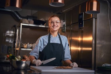 Gülümseyen kadın şef, mutfak masasının yanında duruyor, bıçak tutuyor, önlük ve gömlek giyiyor, kameraya poz veriyor, realite şovu gibi.