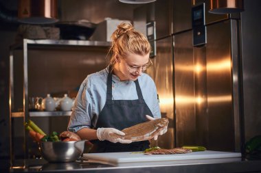 Curious female chef standing in a kitchen next to cutting board, wearing apron and shirt, posing for the camera, reality show look
