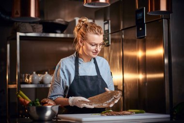 Interested female chef standing in a kitchen next to cutting board, holding a fish, wearing apron and shirt, posing for the camera, reality show look