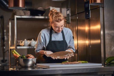 Interested female chef standing in a kitchen next to cutting board, holding a fish, wearing apron and shirt, posing for the camera, reality show look