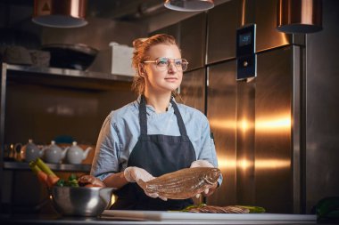 Interested female chef standing in a kitchen next to cutting board, holding a fish, wearing apron and shirt, posing for the camera, reality show look