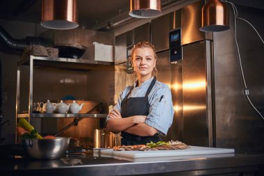 European female chef standing in a kitchen next to cutting board, hands crossed, wearing apron and shirt, posing for the camera, reality show look