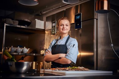 Confident female chef standing in a kitchen next to cutting board, with arms crossed, wearing apron and shirt, posing for the camera, reality show look