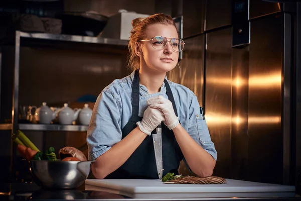 レストランのキッチンに立つ思考と平和的な女性シェフ、カメラで辛抱強く見てまな板に対してナイフを保持. — ストック写真