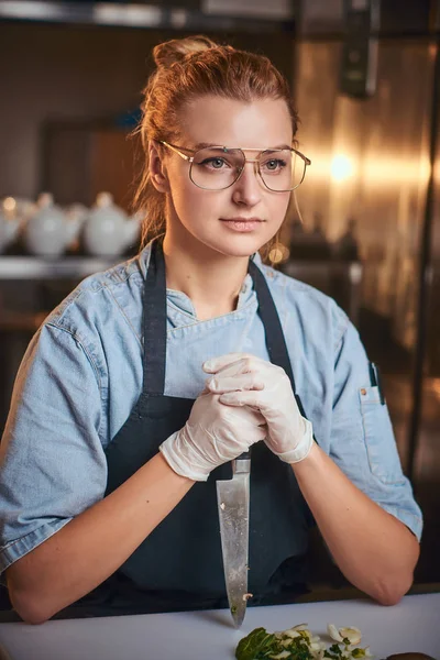 Pensando e tranquillo chef femminile in piedi in cucina ristorante, tenendo un coltello contro tagliere guardando pazientemente sulla macchina fotografica . — Foto Stock