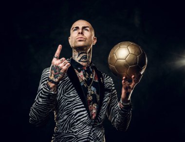 Karanlık bir stüdyoda fotoğraf çekimi için poz veren ve altın bir futbol topu tutan cesur erkek manken.