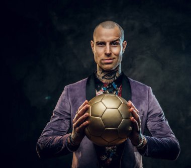 Karanlık bir stüdyoda smokin giyen ve elinde altın bir futbol topu tutan şık dövmeli erkek manken. Kamerada sırıtırken, yakın plan.
