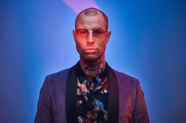 Sakin dövmeli, kel, modaya uygun bir erkek model. Stüdyoda fotoğraf çekimi için poz veriyor. Gri smokin ve gözlük takıyor.