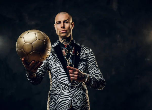 Смелый татуированный мужчина-модель позирует для фотосессии в темной студии и держит золотой футбольный мяч, будучи серьезным — стоковое фото