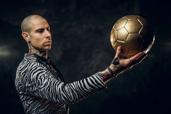 Odważny tatuaż mężczyzna model pozowanie dla sesji zdjęciowej w ciemnym studio i trzymając złoty piłka nożna patrząc na to — Zdjęcie stockowe