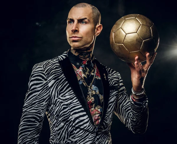 Modèle masculin tatoué audacieux posant pour photoshoot dans un studio sombre et tenant un ballon de football doré tout en regardant loin — Photo