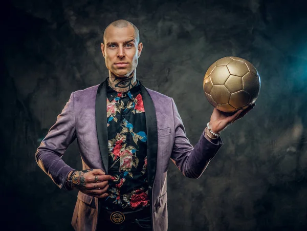 Elegante modelo masculino tatuado con esmoquin posando para una sesión de fotos en un estudio oscuro y sosteniendo una pelota de fútbol dorado mientras se ve confiado en la cámara —  Fotos de Stock