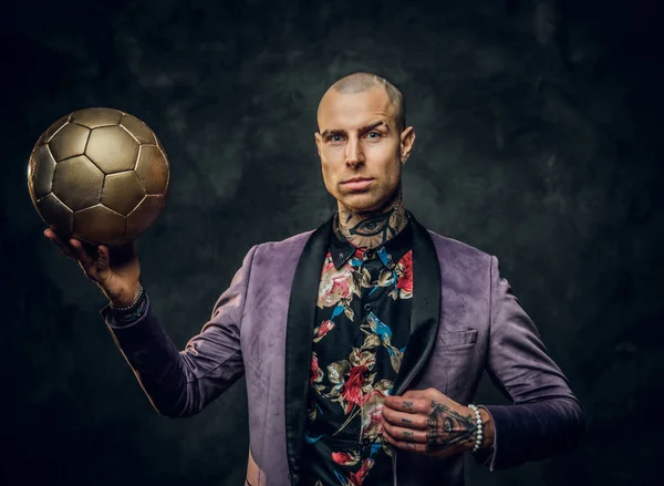 Atrevido modelo masculino tatuado con esmoquin posando para una sesión de fotos en un estudio oscuro y sosteniendo una pelota de fútbol dorado mientras mira a la cámara —  Fotos de Stock