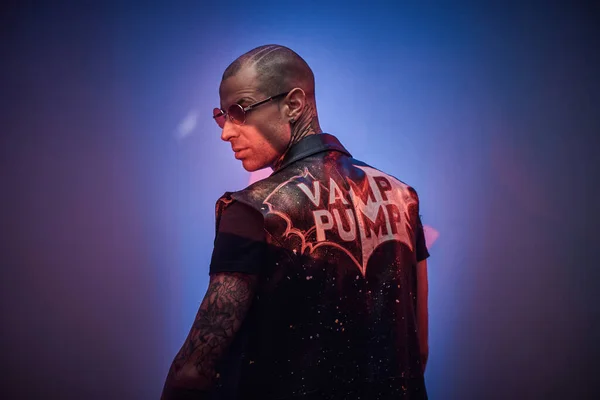 Brutal tattooed, bald fashionable male model posing in a studio for the photoshoot vogue style, looking from behind