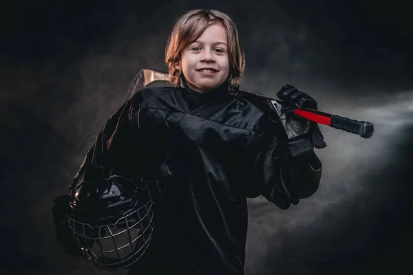 Young hockey player posing in uniform for a photoshot in a studio while holding his helmet and hockey stick — Stock Photo, Image